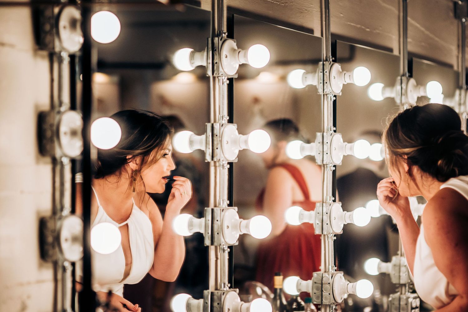 Person applying makeup in a dressing room mirror surrounded by bright vanity lights.