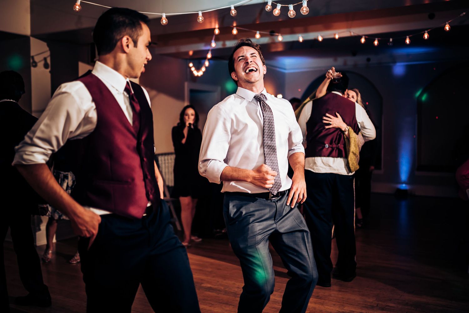 Men dancing and celebrating at a lively indoor reception with festive lighting.