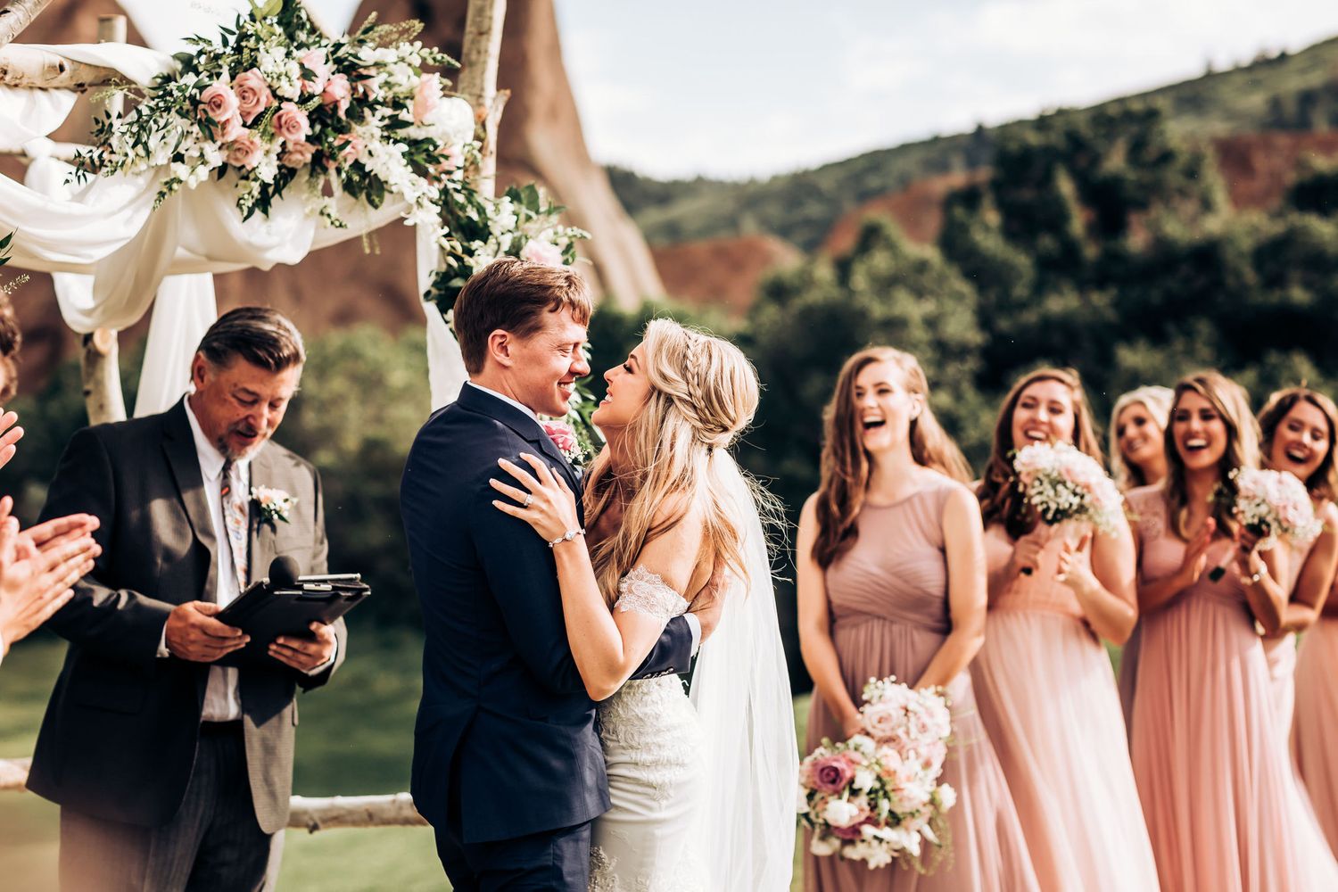 Wedding party celebrates during ceremony with bridesmaids in blush pink dresses under a decorated floral arch.