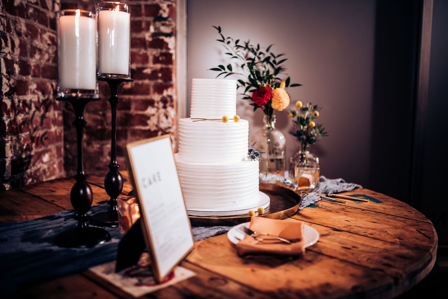 Elegant white wedding cake with fresh floral decorations displayed on rustic wooden table with candlelight.