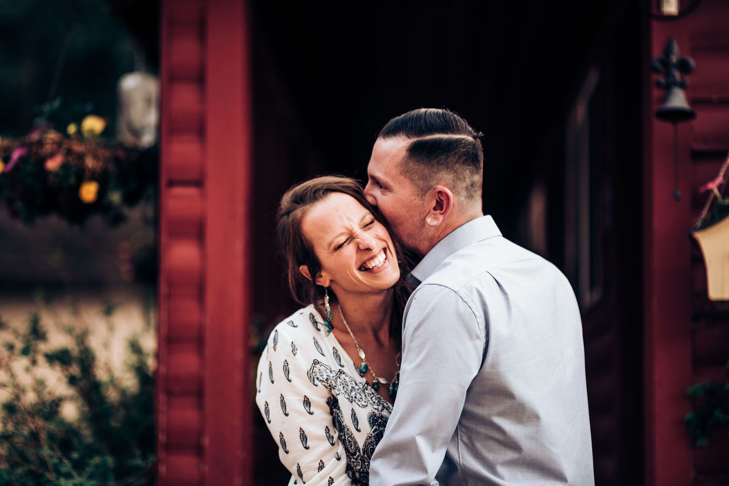 Couple sharing laughter and embrace against rustic red building background.