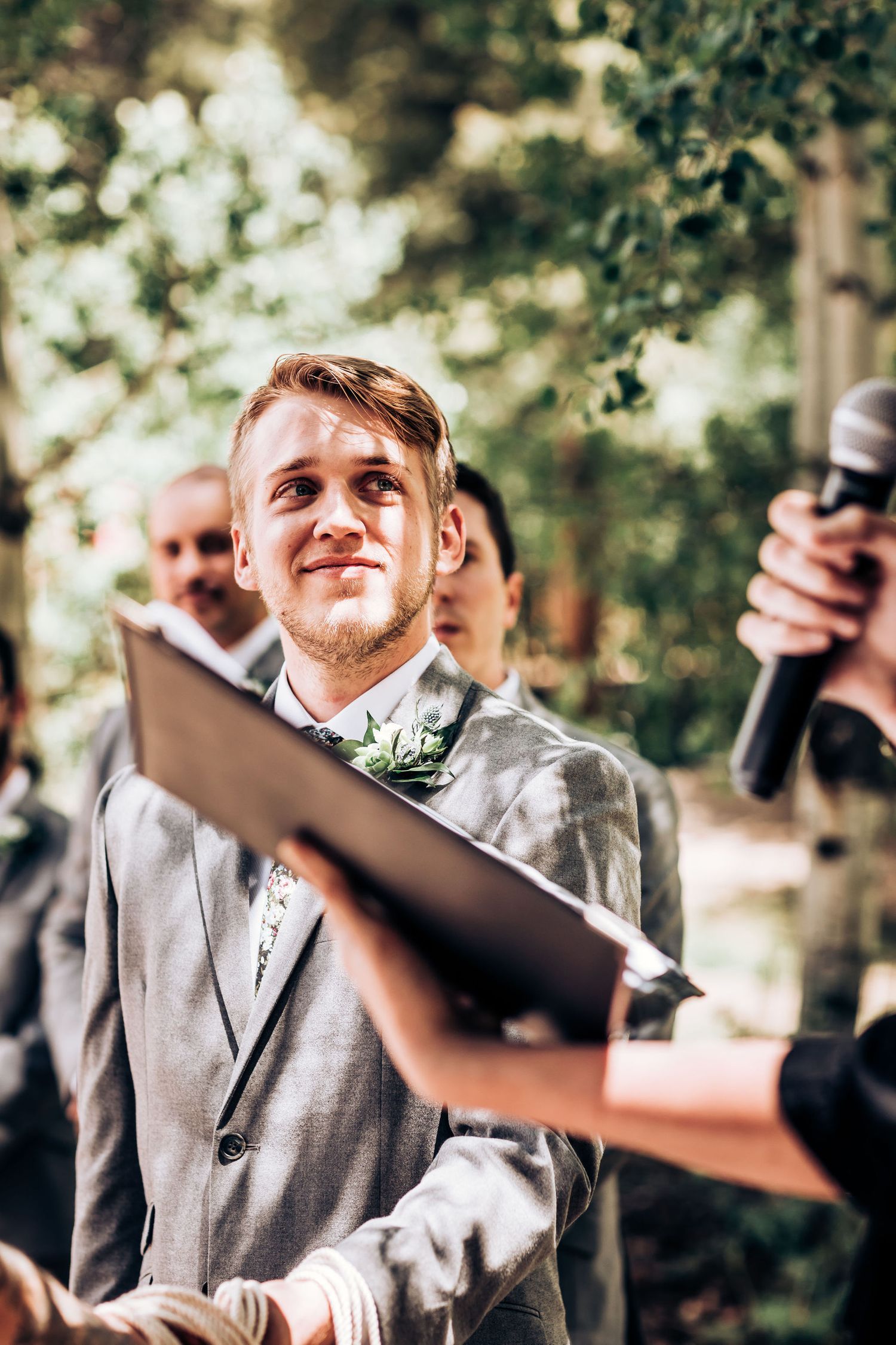 A groom in a light grey suit smiles during an outdoor forest wedding ceremony.