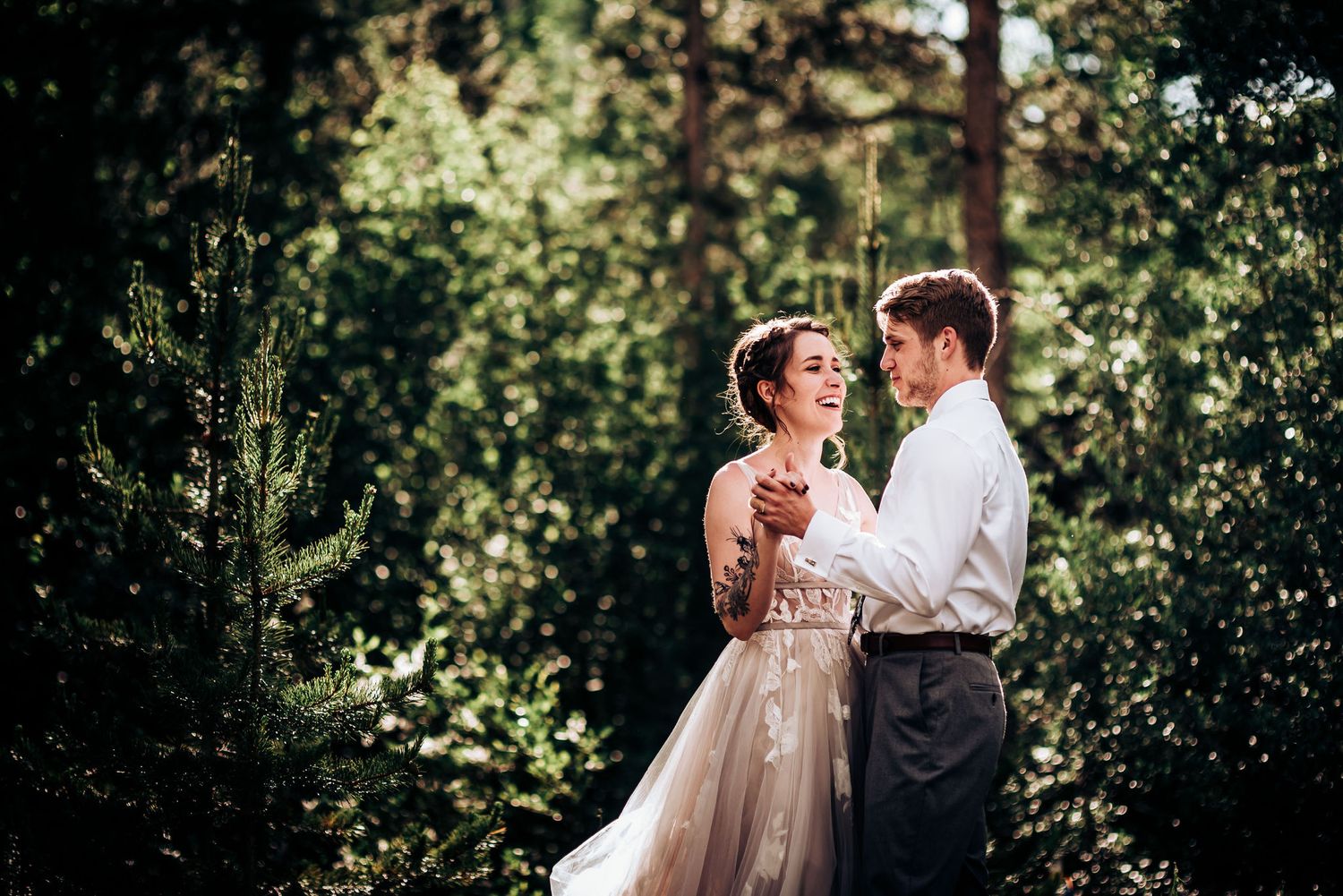An intimate embrace captured in a sun-dappled forest setting with pine trees in the background.