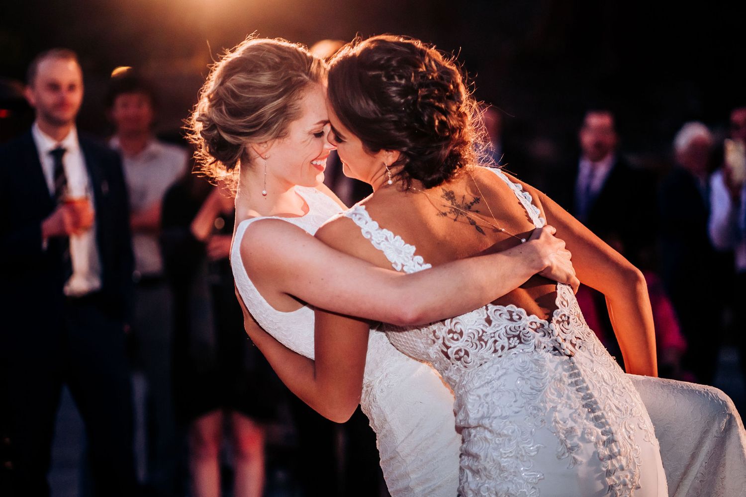 Two brides in white lace wedding dresses embrace warmly on the dance floor during their wedding reception.