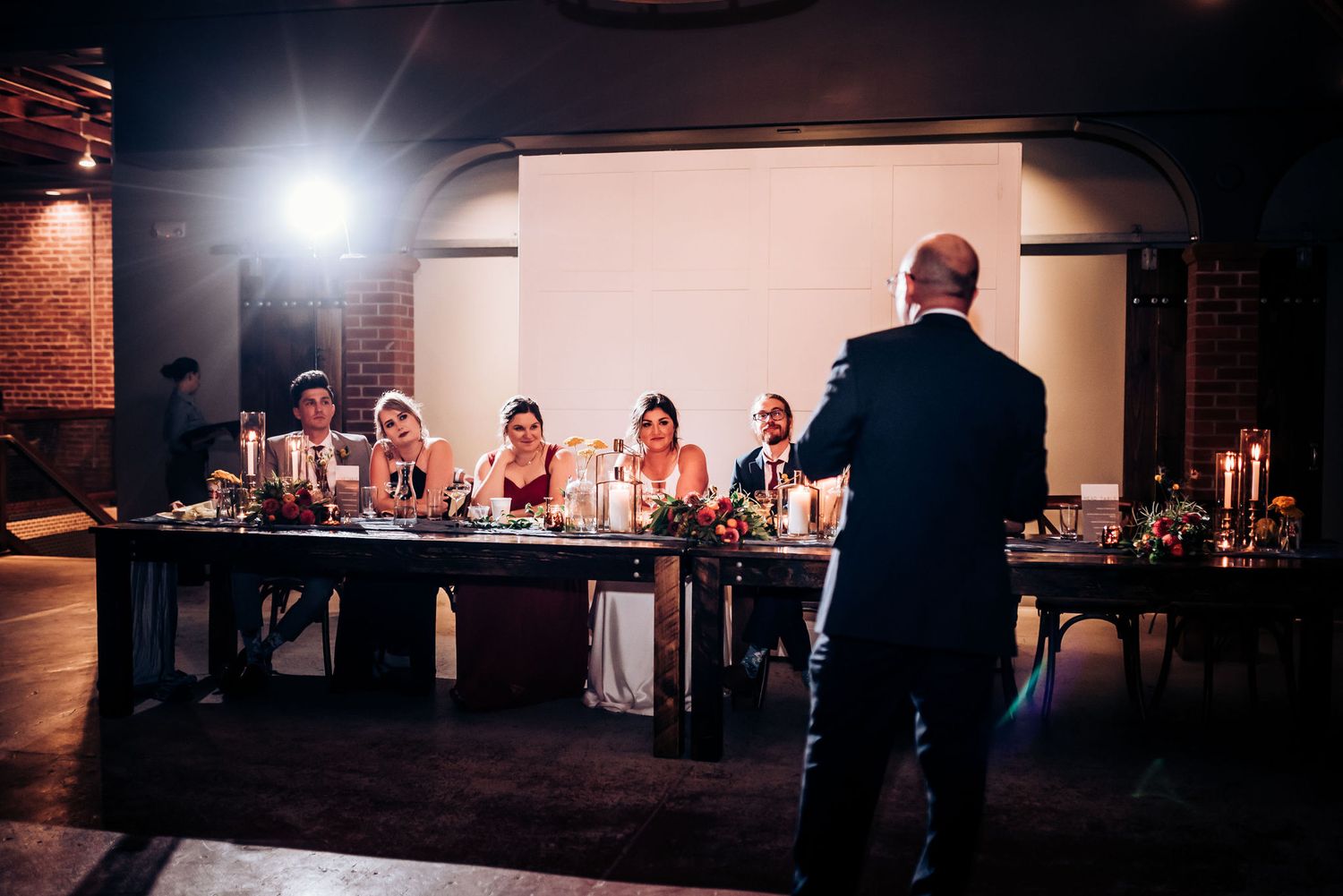 Event speaker addressing attendees seated at decorated tables in an industrial-style venue.
