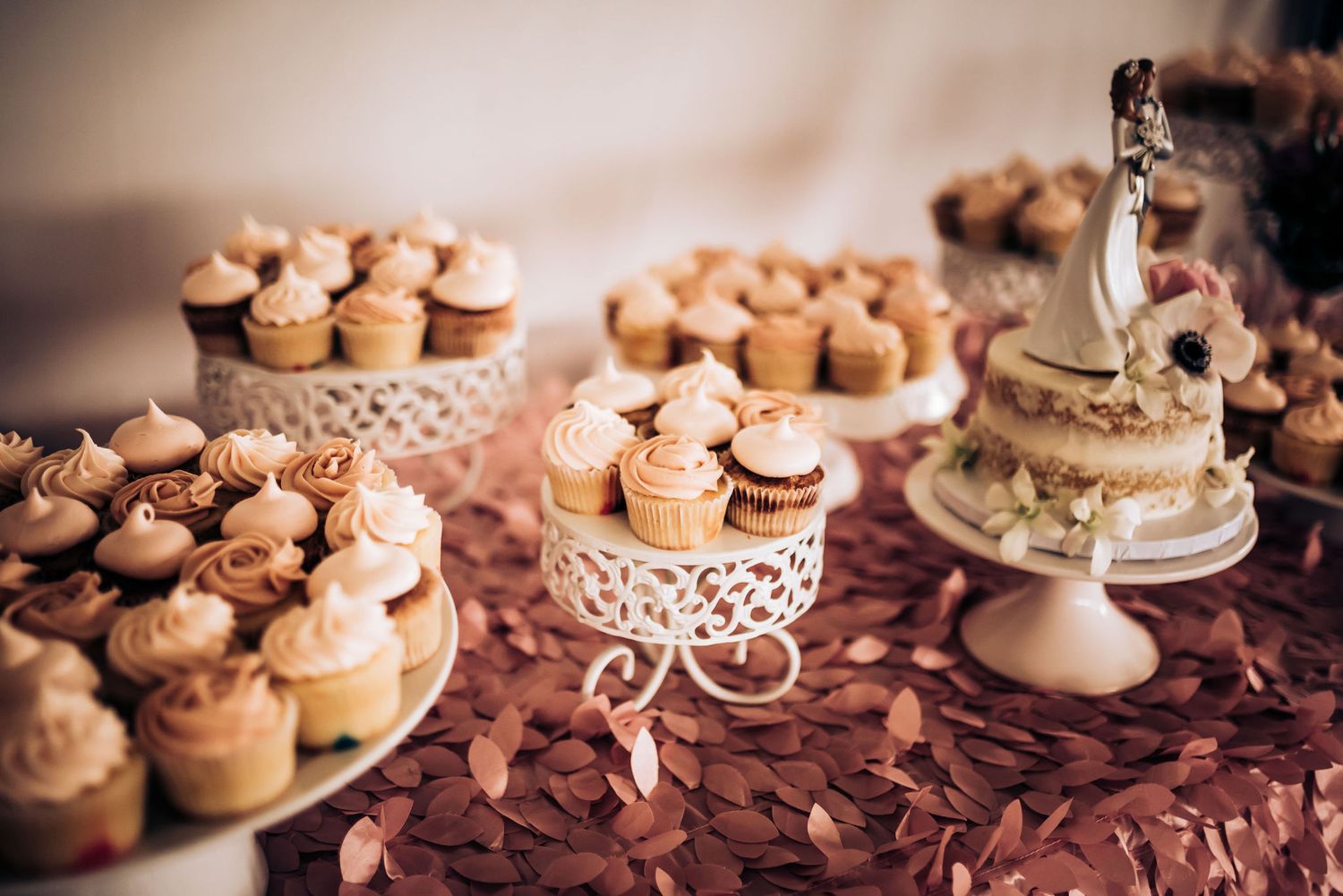 Multiple tiers of cupcakes displayed on ornate white cake stands against a textured burgundy backdrop.
