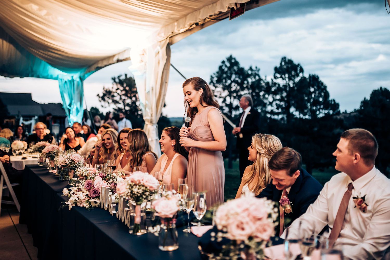 Wedding party members listening to toasts at an elegant outdoor reception under a tent with floral centerpieces.