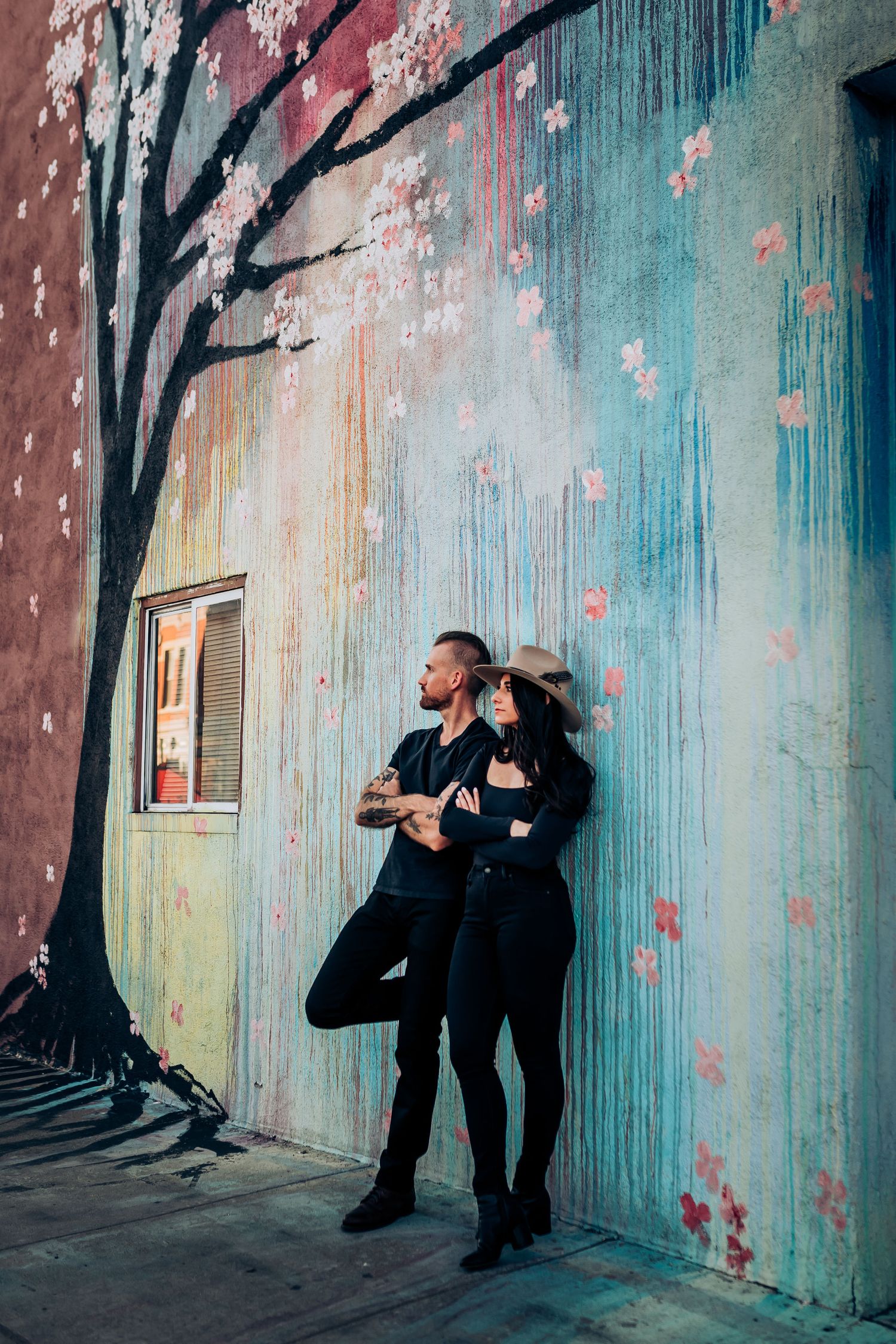 A couple poses against a colorful mural wall with painted cherry blossoms and a small window.