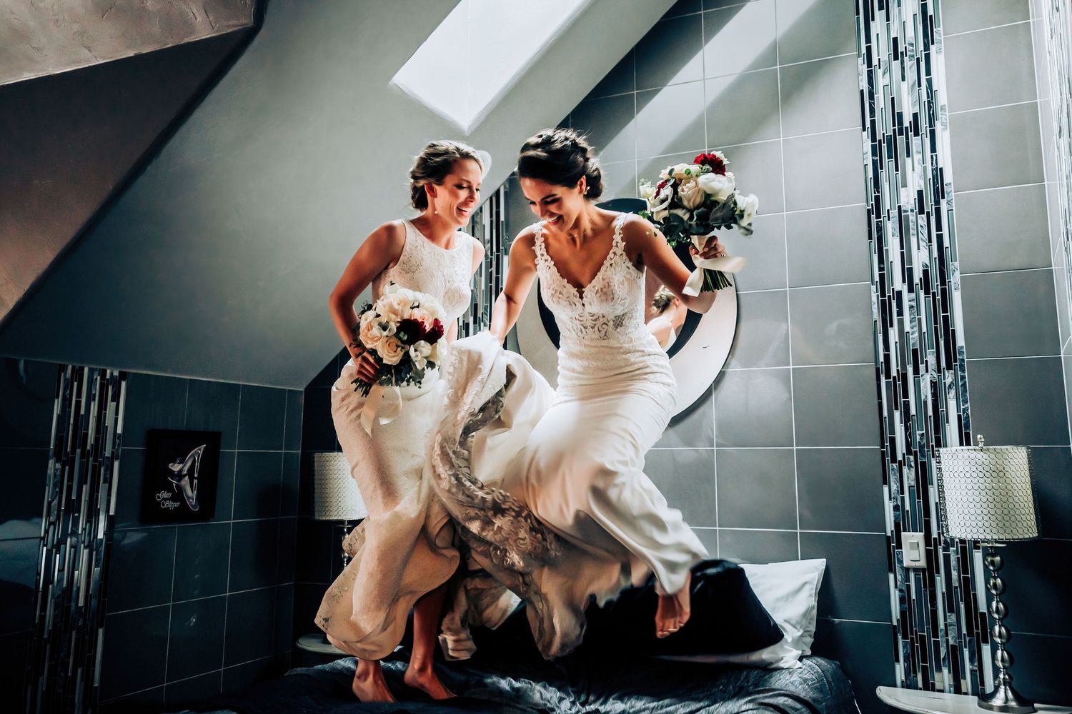 Wedding party members in white dresses jumping playfully in a modern hotel room.