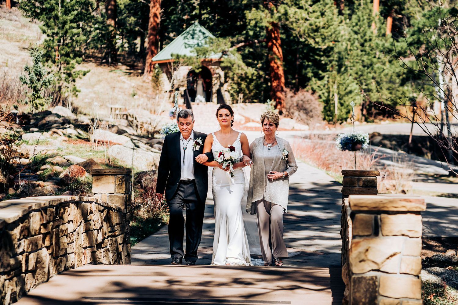Wedding processional walking down stone-lined outdoor pathway surrounded by pine trees.