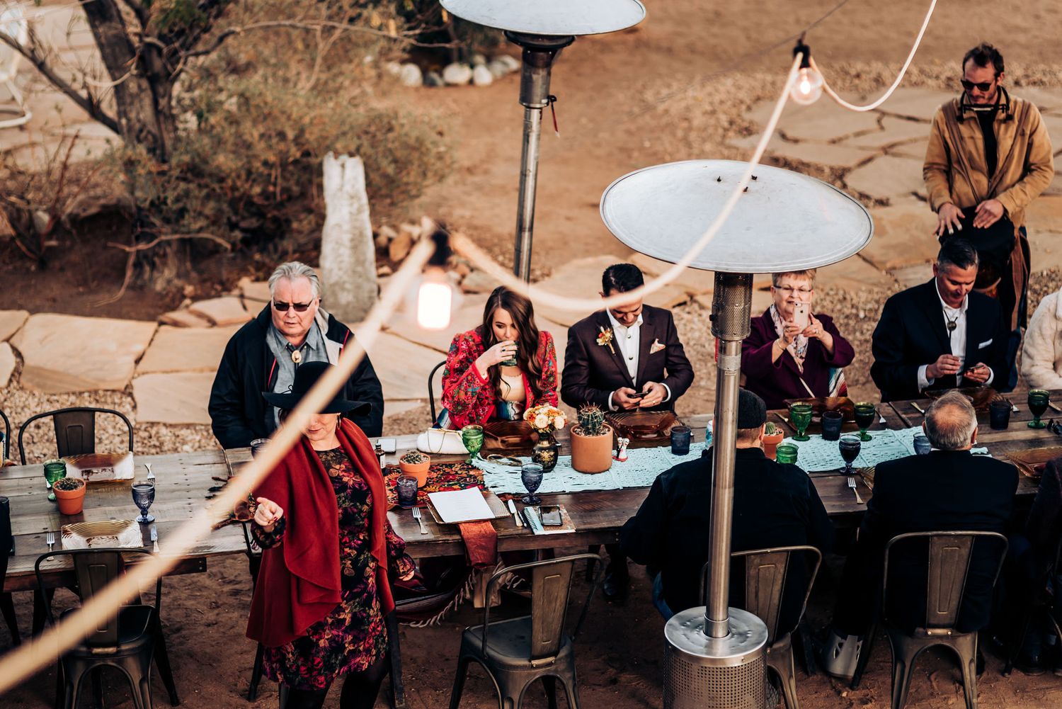 Guests seated at long tables under heating lamps at an outdoor desert reception.