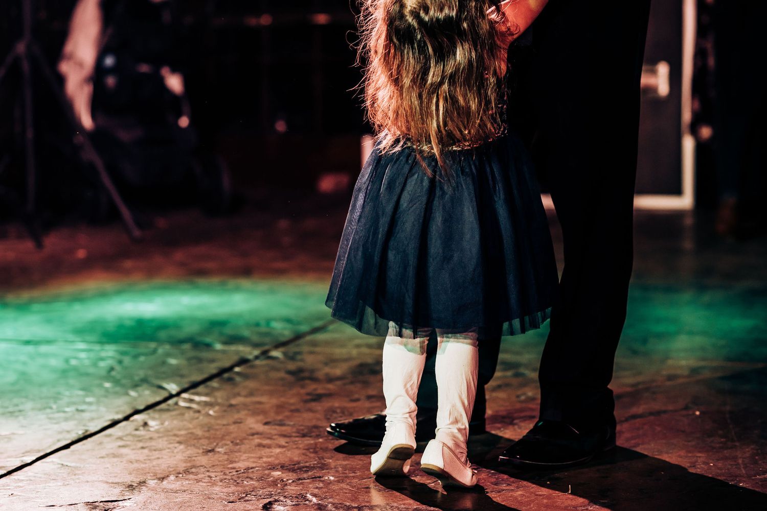 A small child in a navy tulle dress stands on an adult's feet on a colorfully lit dance floor.
