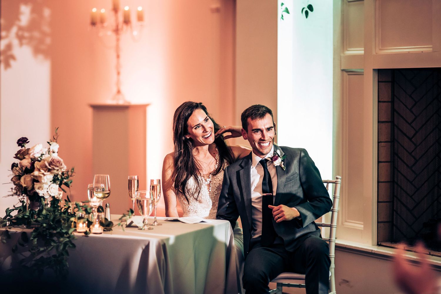 A couple sits together at an elegant reception table with floral arrangements in a warmly lit venue.
