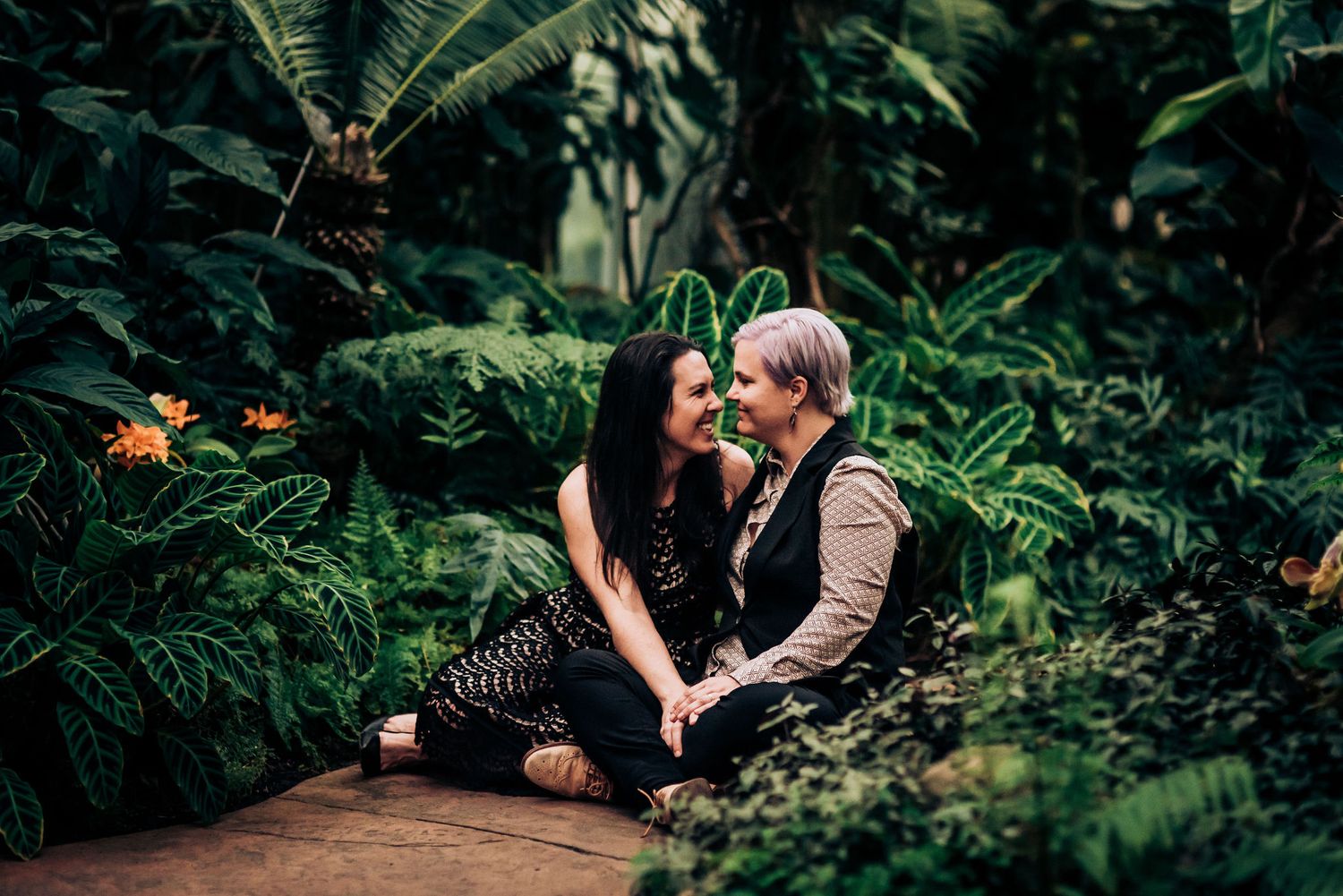 Two people share an intimate moment while sitting on a wooden pathway in a lush garden.