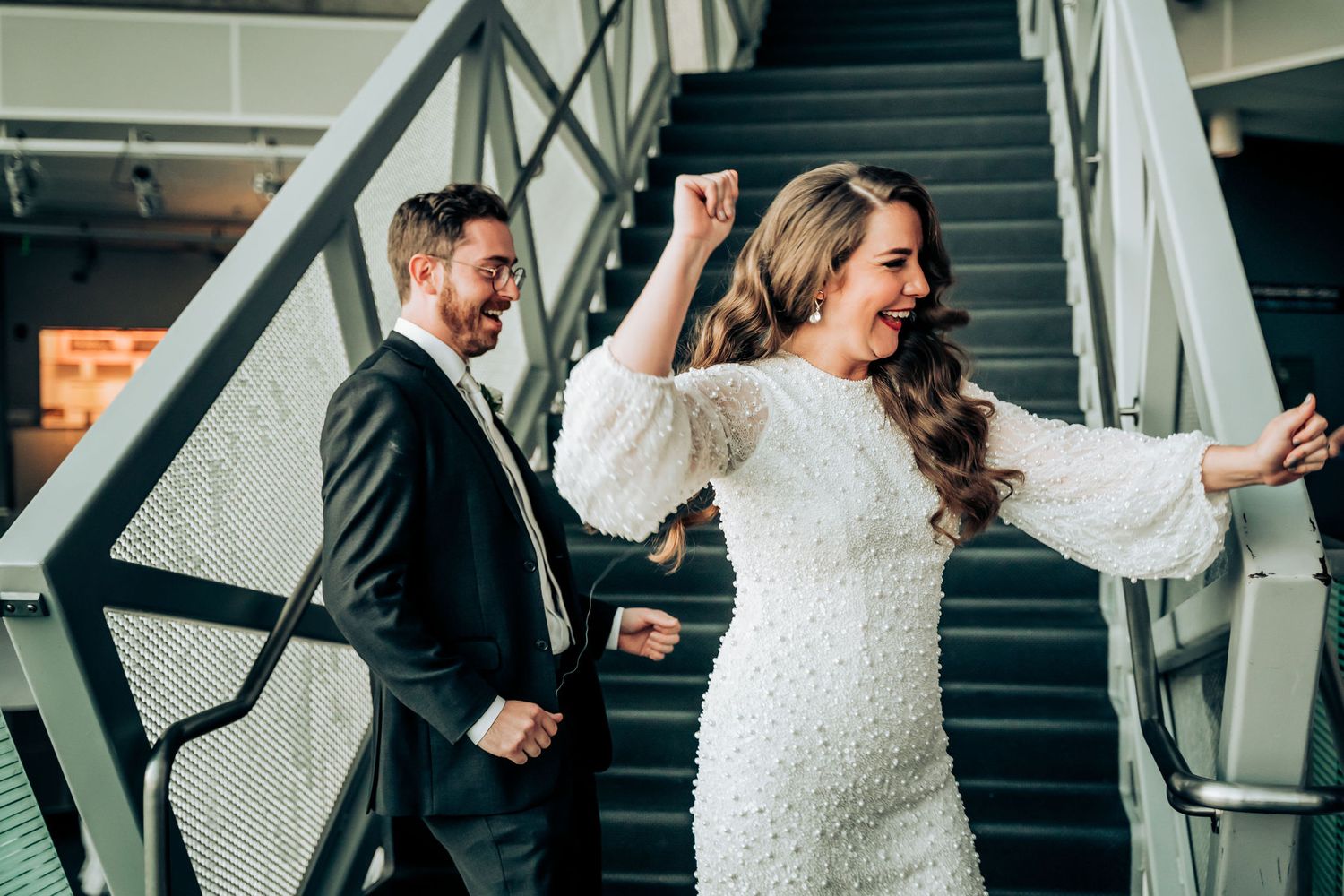 A couple celebrating on a modern staircase, with the bride in a white dress showing joyful movement.