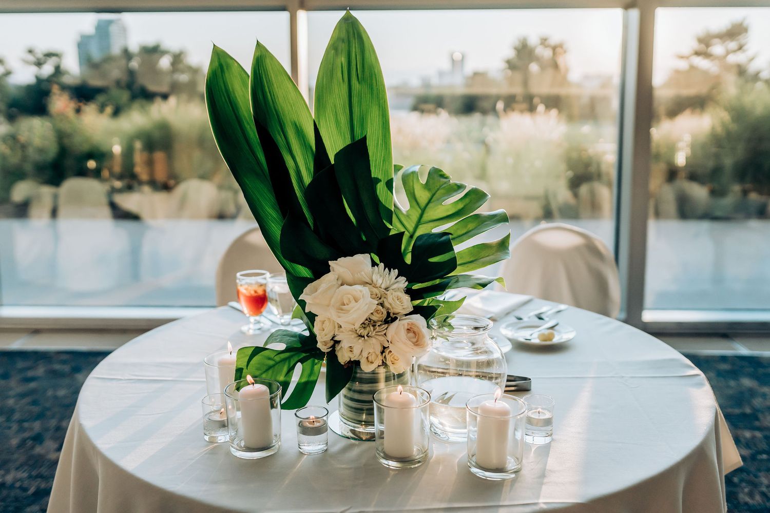 A minimalist table setting with tropical leaf arrangement and glass votives on white linen.
