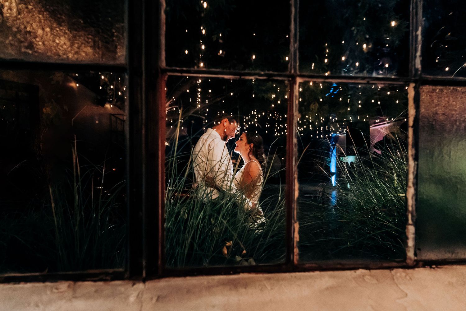 Artistic wedding photograph through a rain-covered window with string lights creating a magical ambiance.