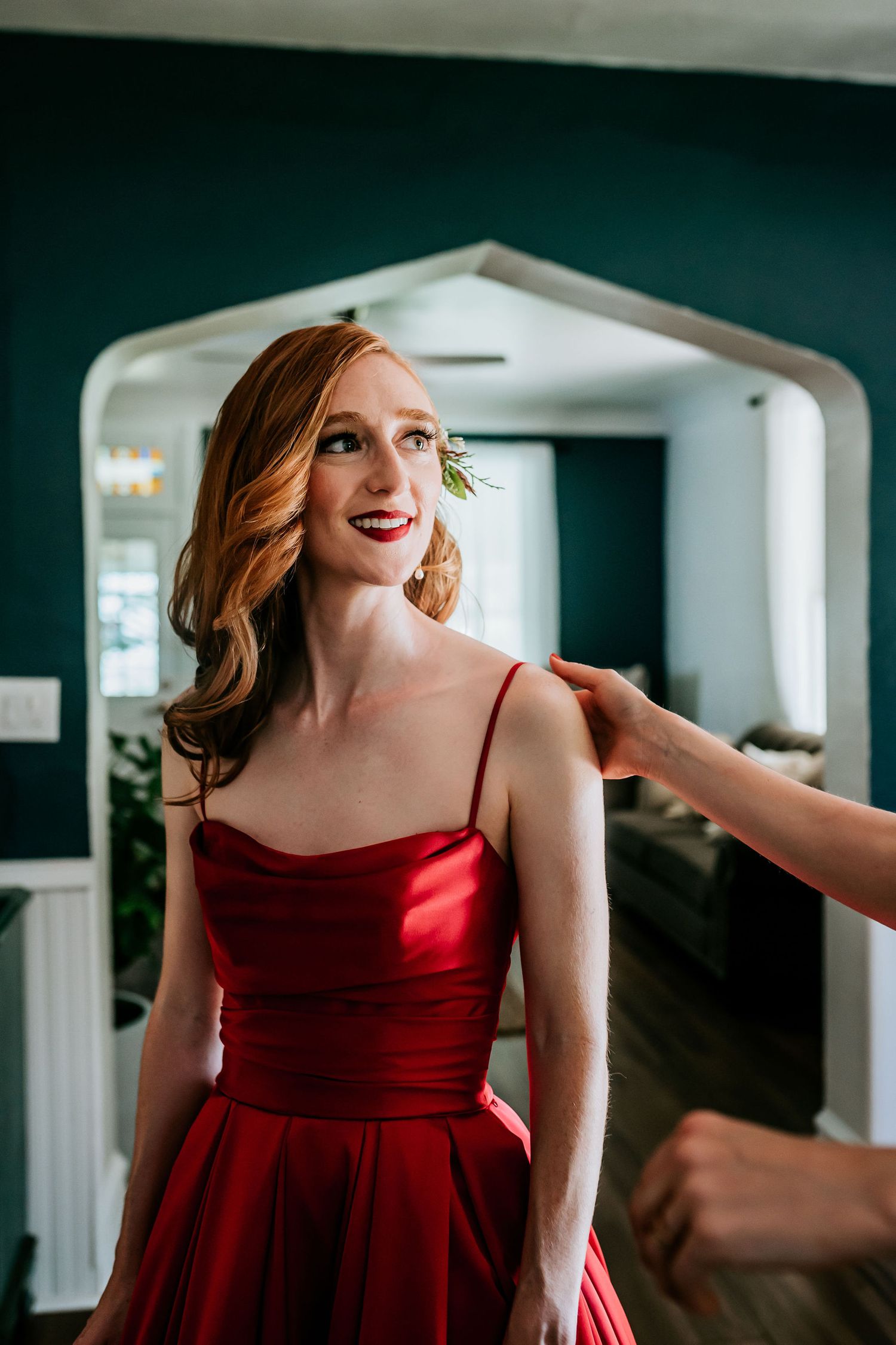 A woman in a stunning red satin dress stands elegantly in an archway while getting ready.