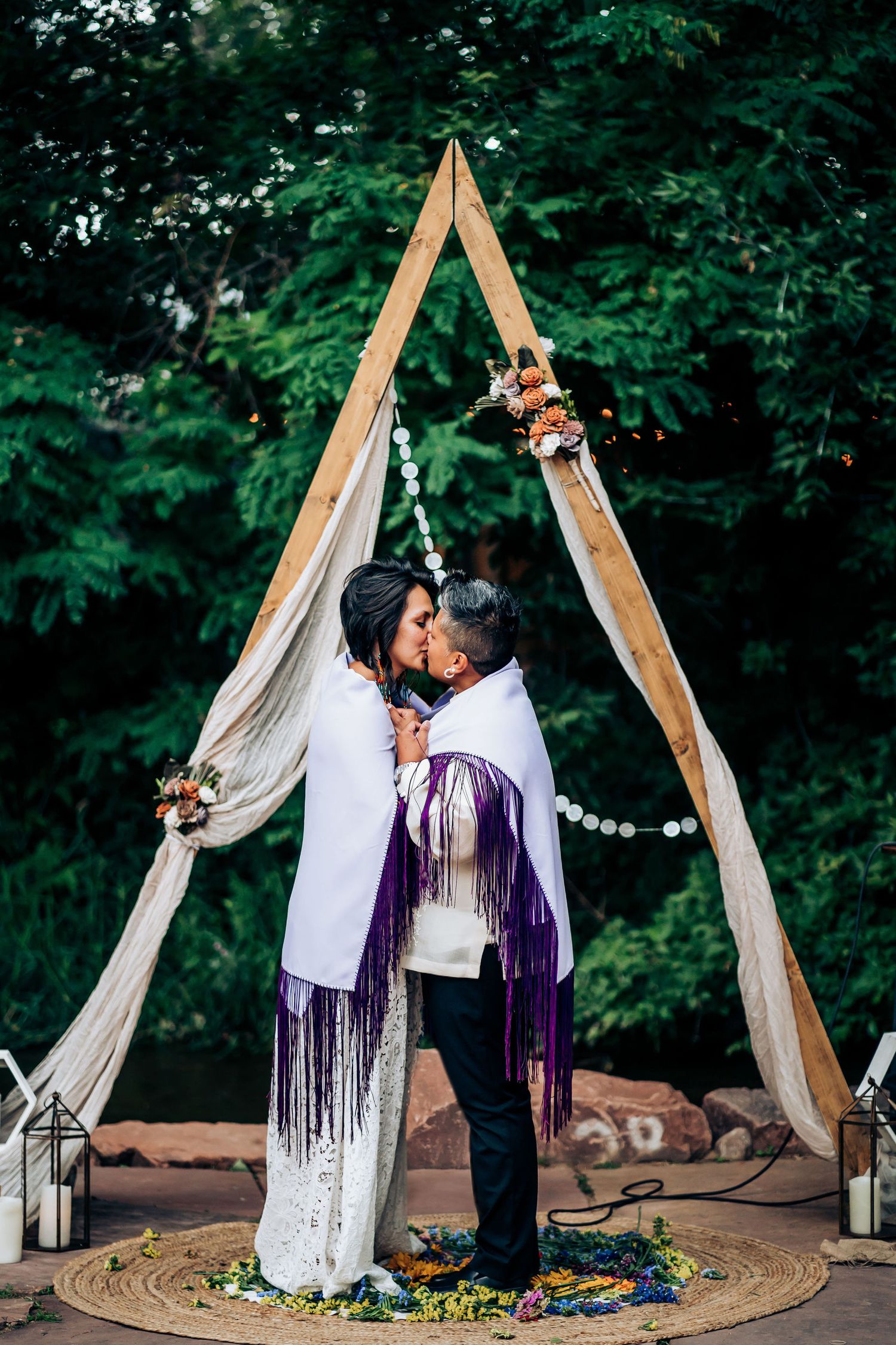 A romantic moment captured under a wooden triangular arch decorated with lights and fabric in a garden setting.