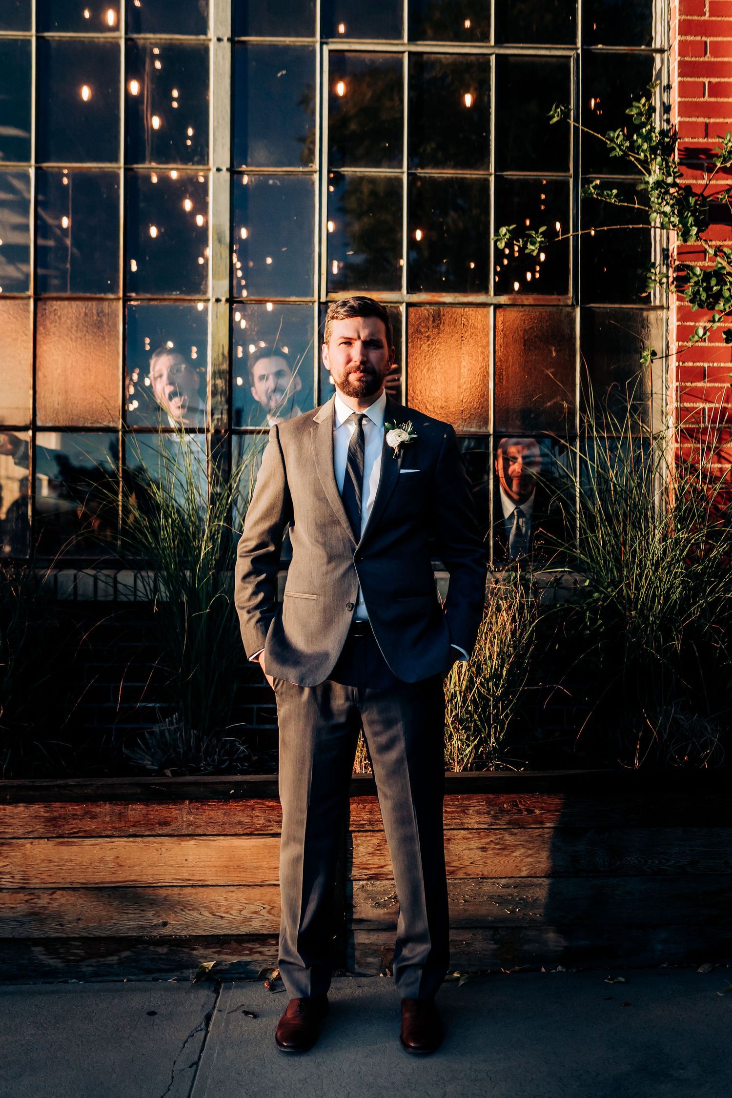 Groom in tailored suit standing against industrial window with string lights.