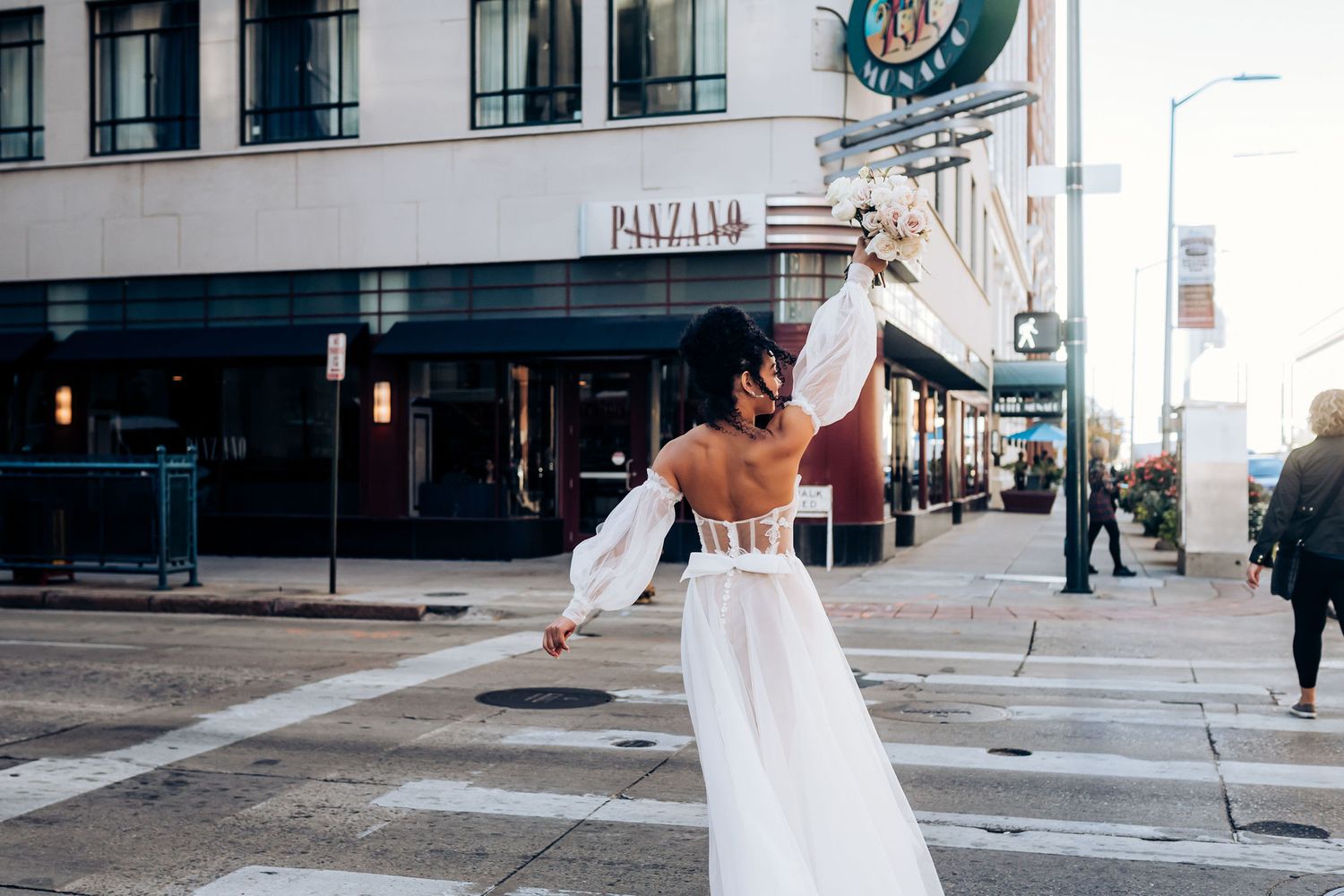 Joyful bride in white dress dancing on city sidewalk with buildings in background.