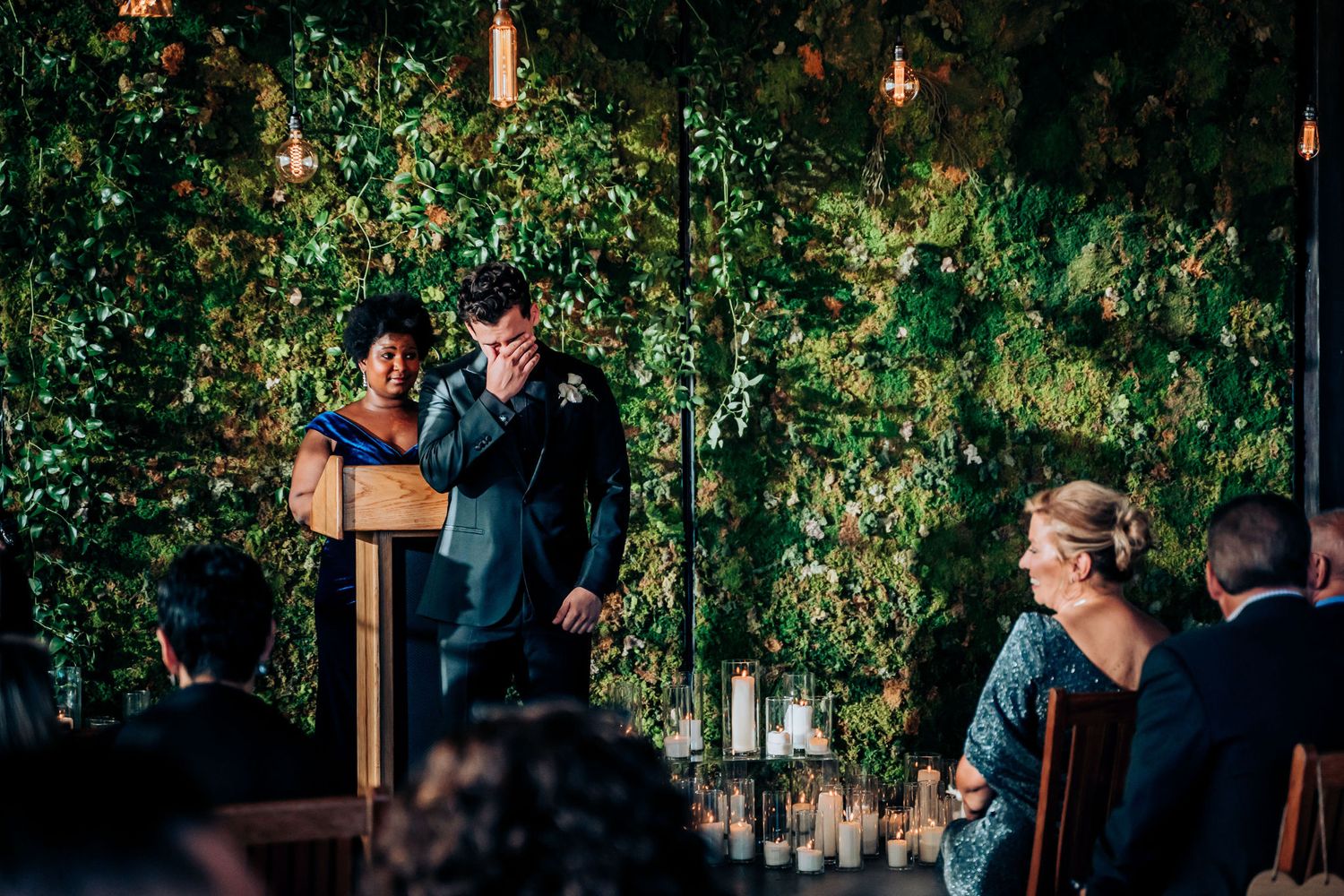 Wedding ceremony in progress with greenery backdrop and hanging lights.