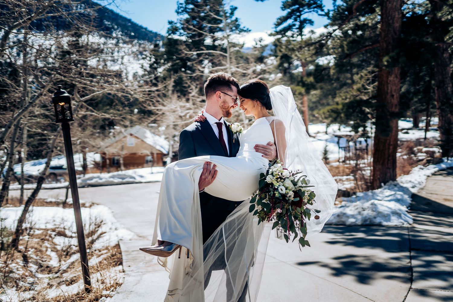 Groom carries bride in a traditional pose against a snowy mountain backdrop.