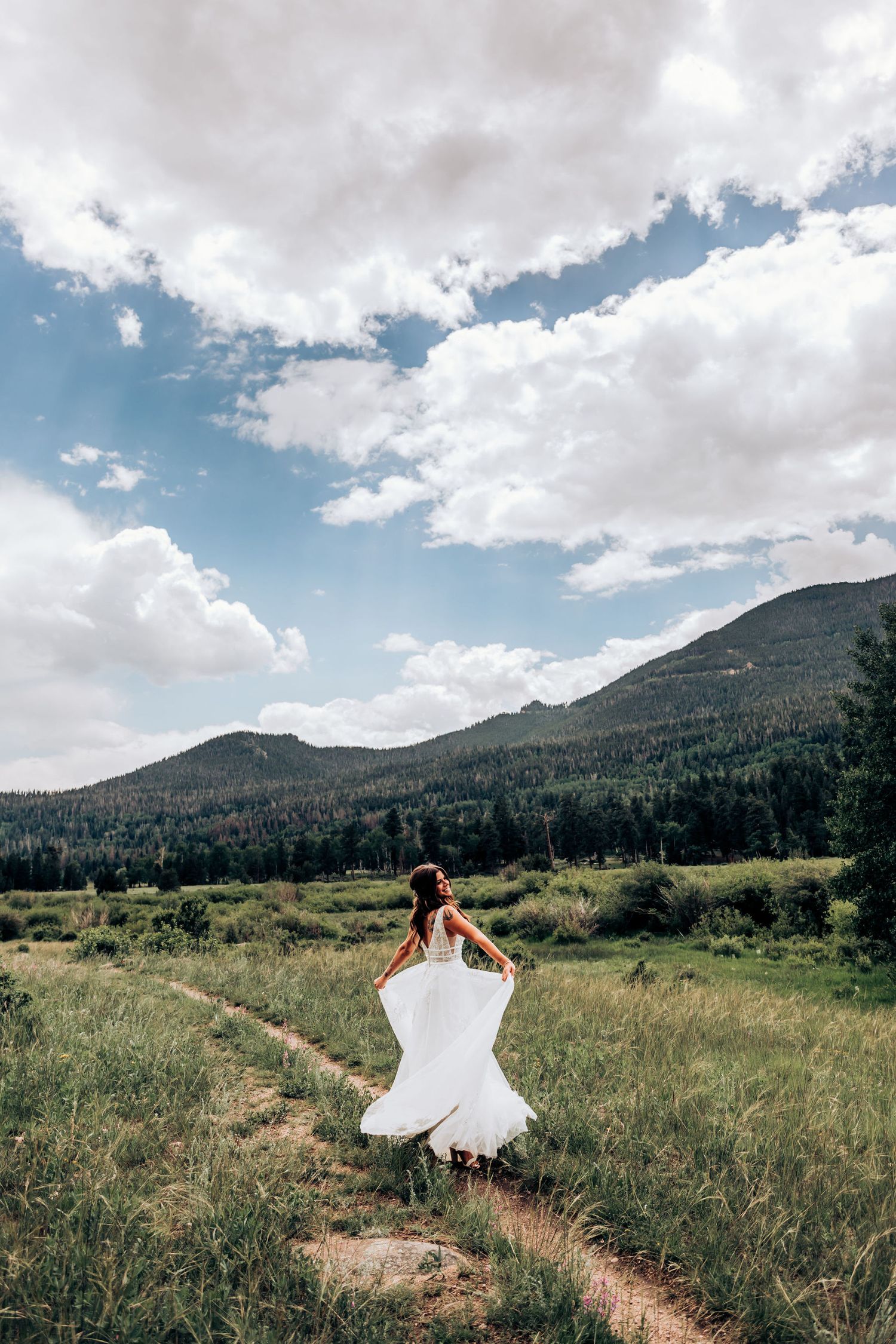 A bride in white dress walks along a mountain trail under dramatic cloudy skies.