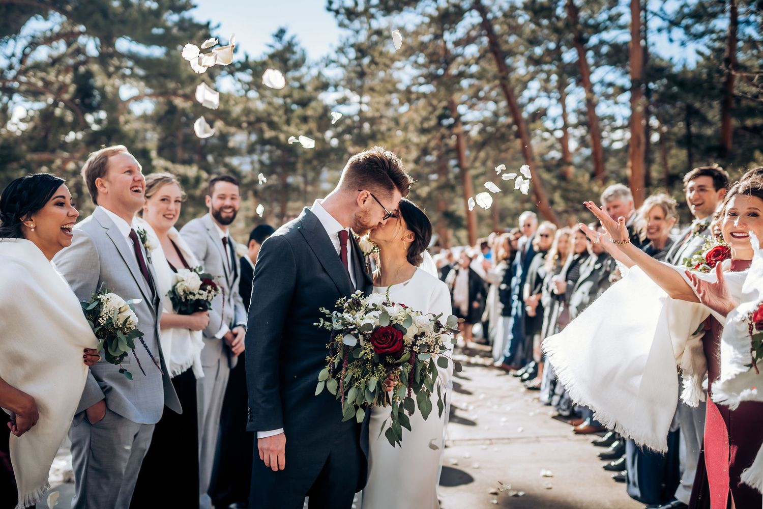 Joyful wedding celebration with guests lined up outdoors among pine trees.