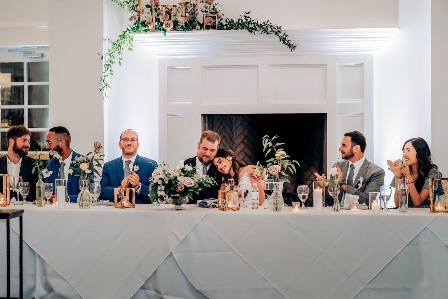 Wedding party seated at long white head table decorated with greenery and candles during reception.