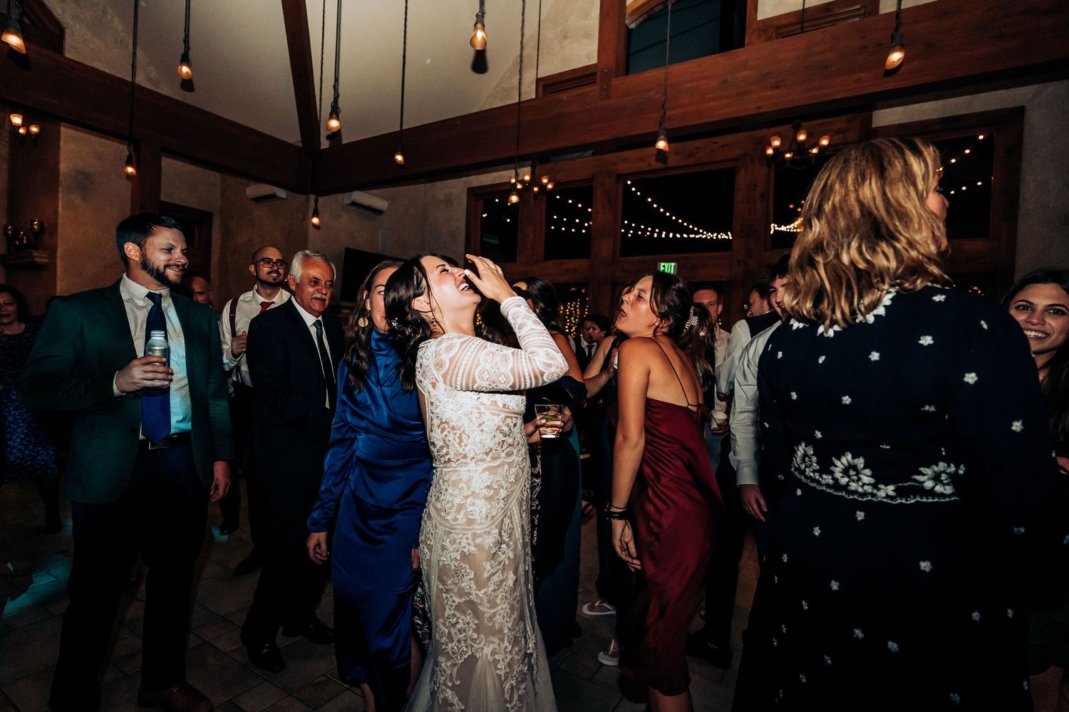 Celebratory scene at a wedding reception with guests dancing beneath rustic wooden beams and warm lighting.