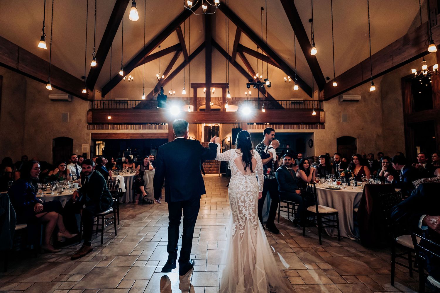 Wedding couple makes a grand entrance into a rustic reception hall with exposed wooden beams and elegant lighting.