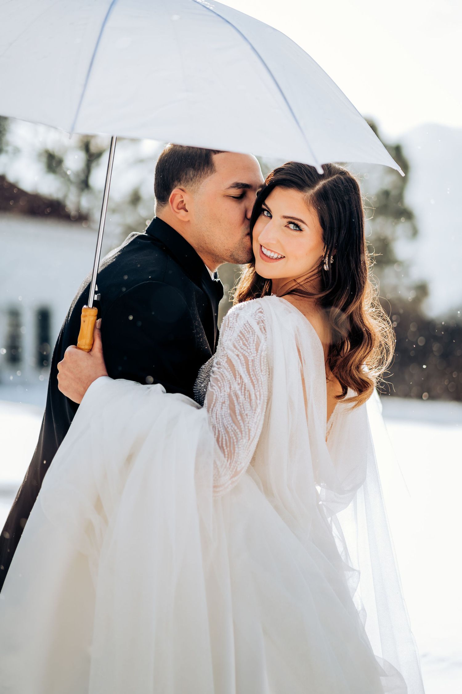 A romantic winter wedding photo showing a couple under a white umbrella in the snow.