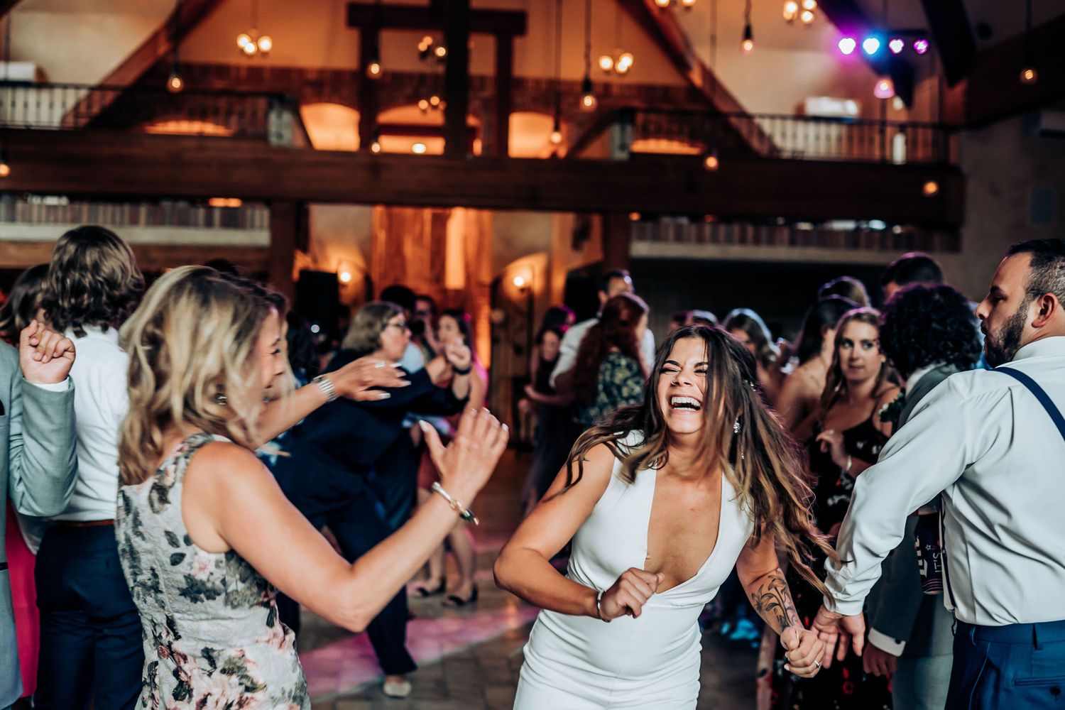 Party guests dance and celebrate on a lively dance floor beneath wooden beams and ambient lighting.