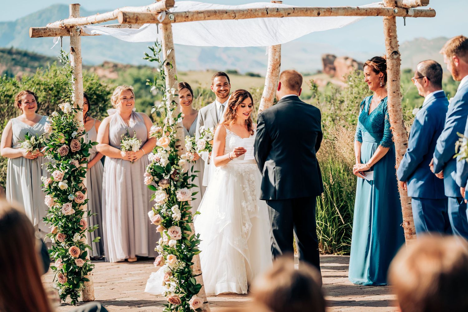Wedding party gathers under decorated wooden arch during mountaintop ceremony.