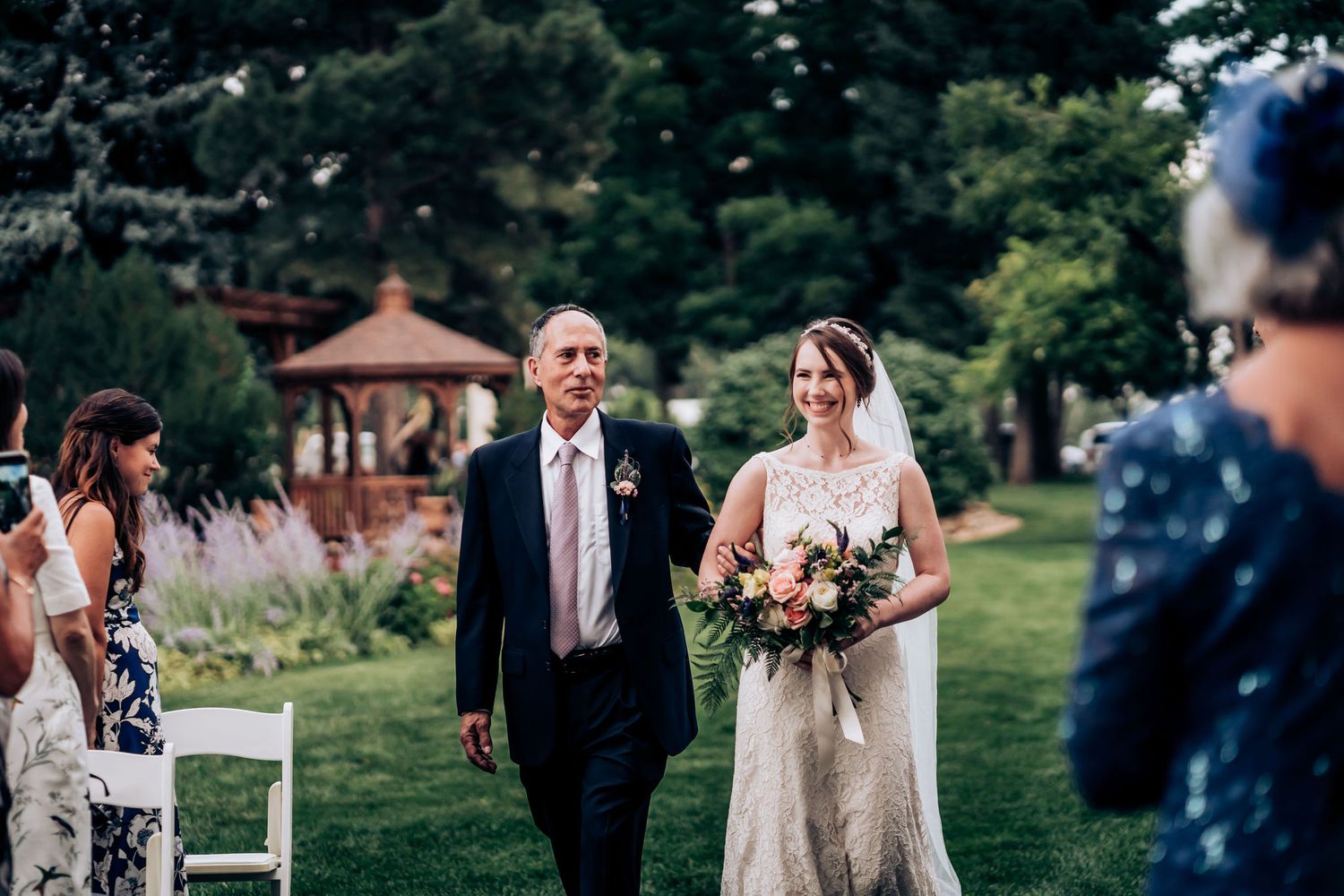 Wedding ceremony processional outdoors with guests seated in white chairs and a garden gazebo in background.