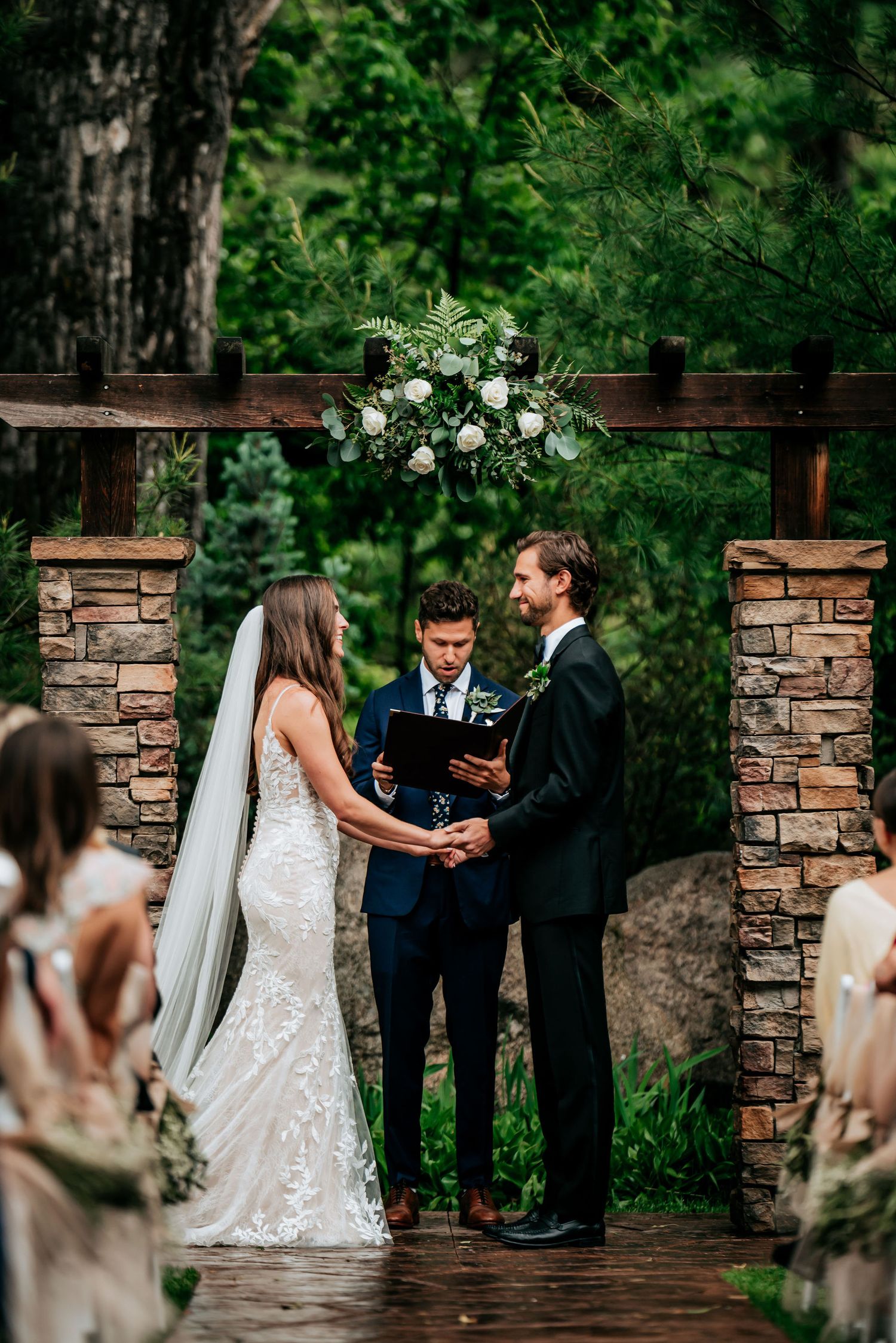 Intimate outdoor wedding ceremony under a floral arch with stone pillars and greenery.