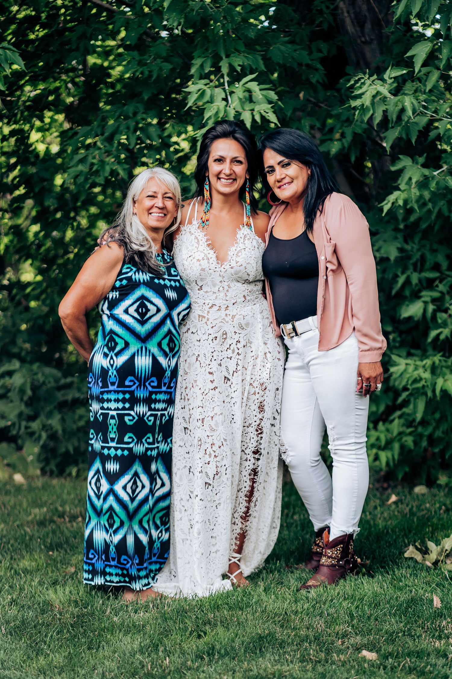 Three women pose together at an outdoor event, with one wearing a white lace wedding dress.