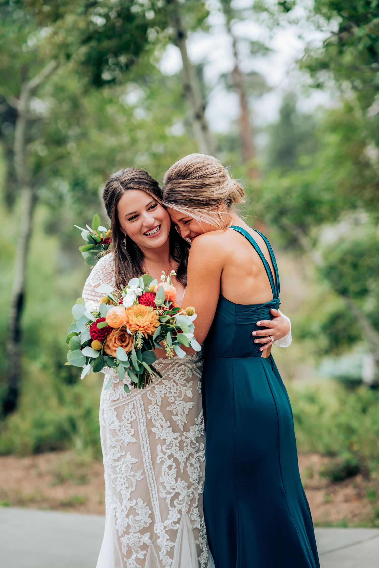 Two wedding attendants share an emotional embrace while holding colorful bouquets outdoors.