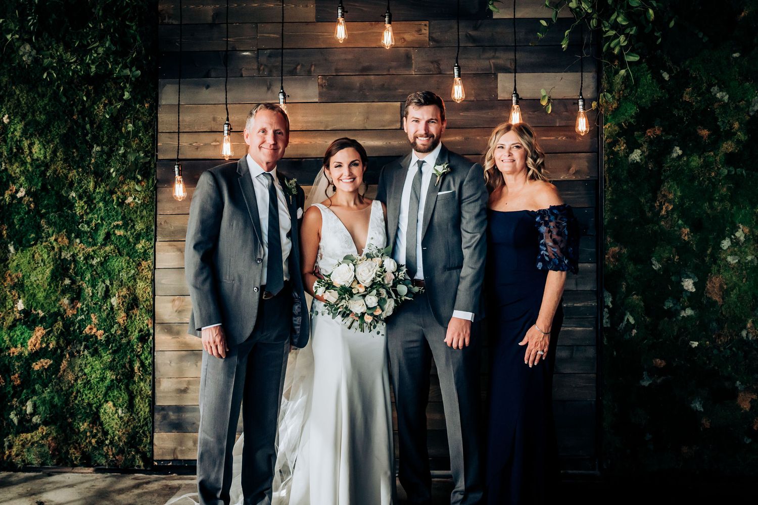 Small wedding group poses against wooden wall decorated with hanging lights and greenery.