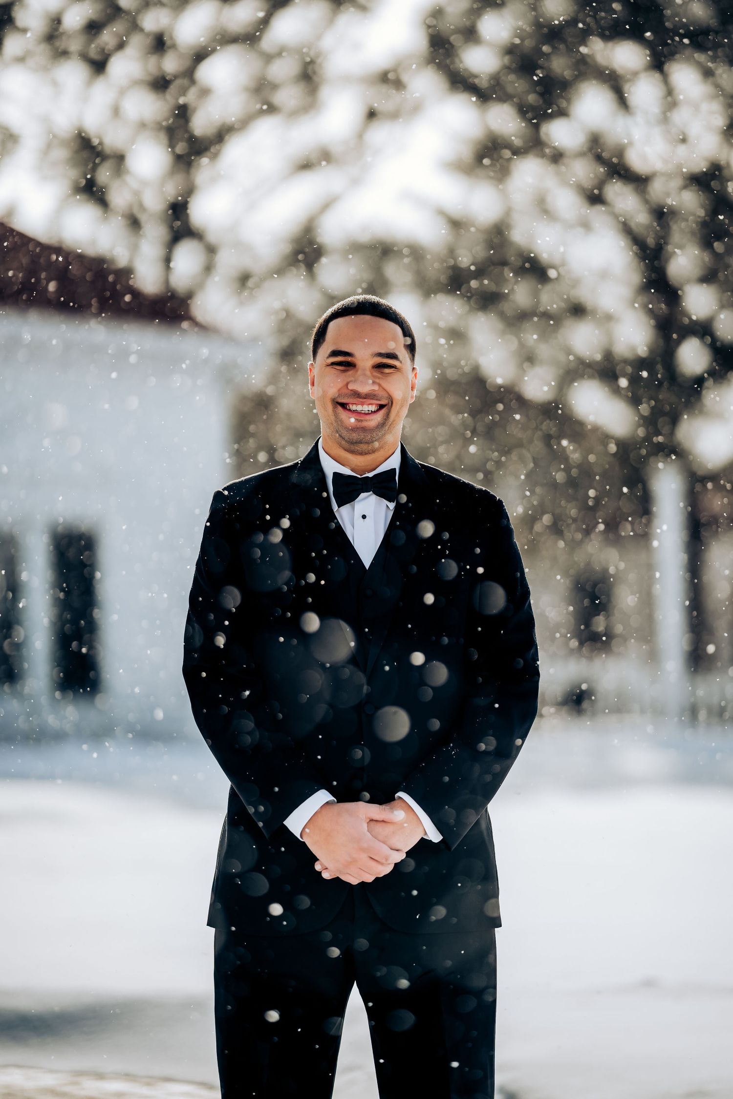 A groom in classic black tuxedo poses in falling snow against winter backdrop.