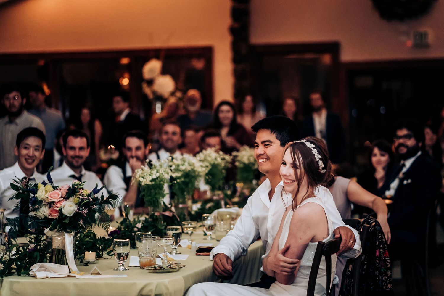 Wedding reception guests seated at decorated tables with white floral centerpieces during a joyous celebration.