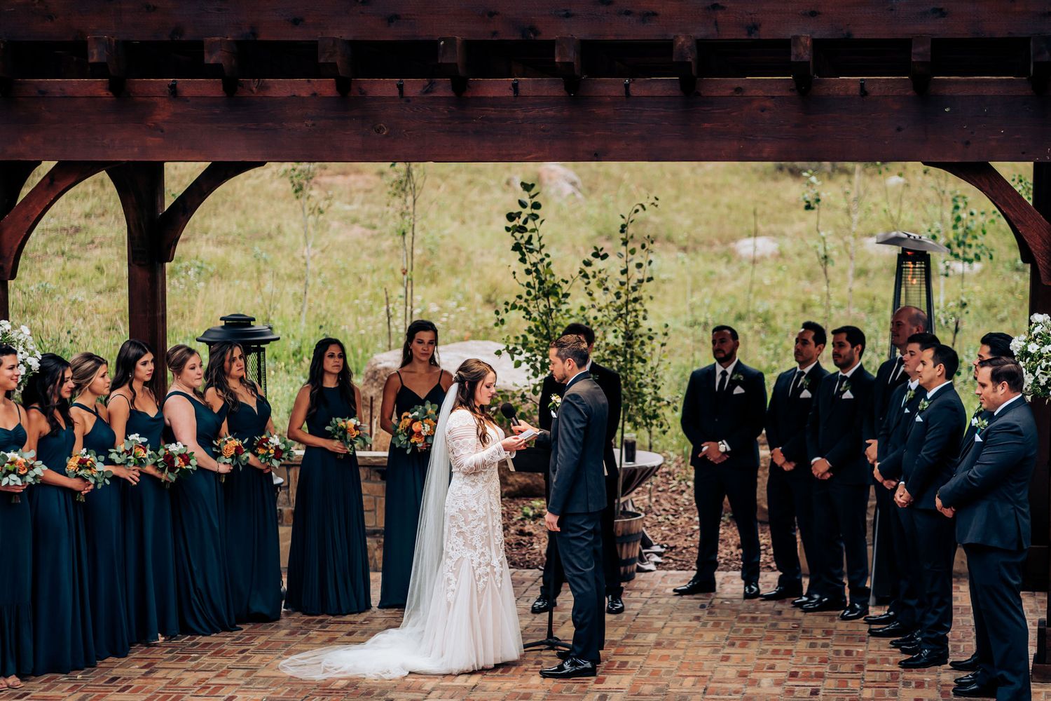 Wedding party in navy blue attire forms a semi-circle around the couple during an outdoor ceremony.