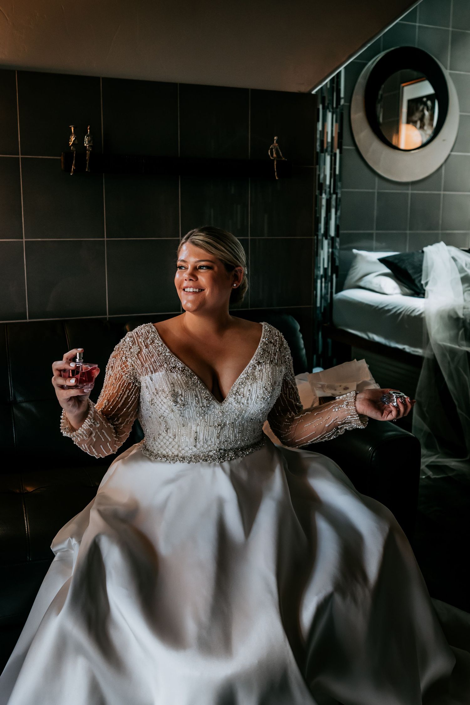 A bride in an elegant beaded gown sits in a modern dark-tiled room holding perfume bottles.