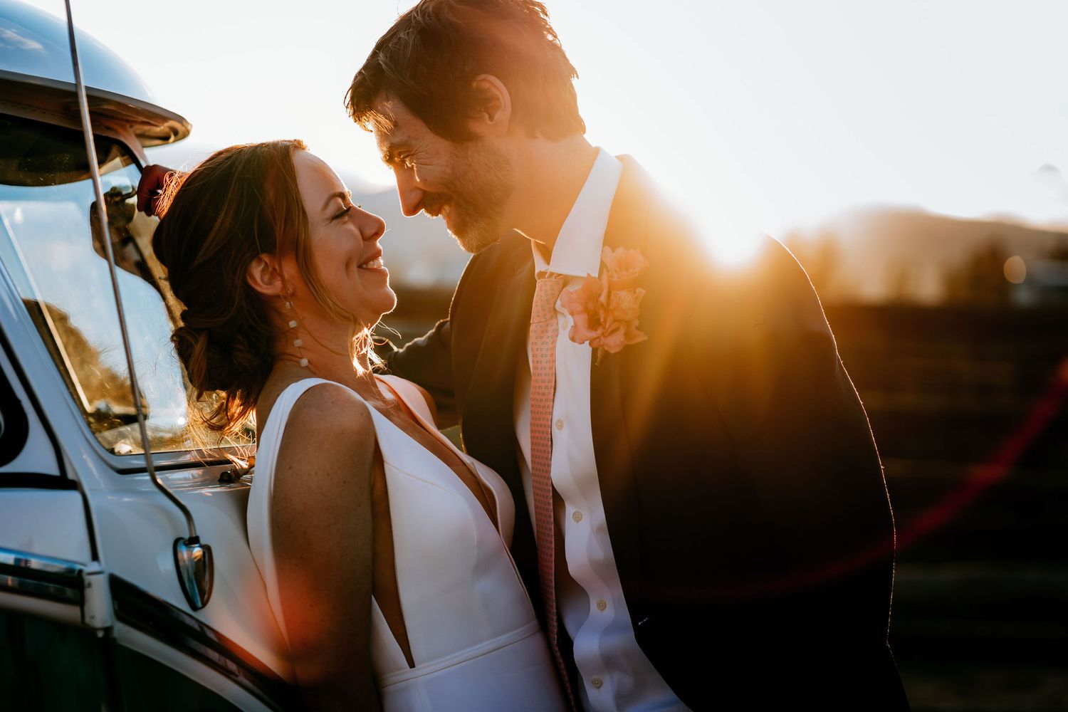 A couple shares an intimate moment at sunset next to a vintage vehicle in romantic wedding attire.