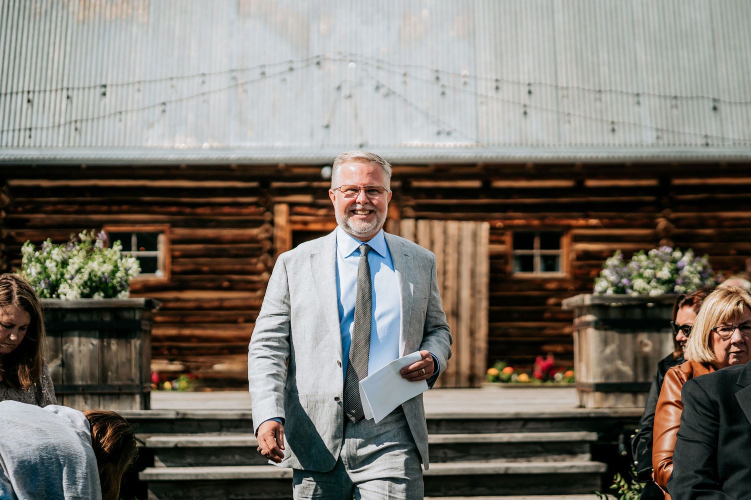 A person in a light gray suit stands on wooden steps outside a rustic log cabin.