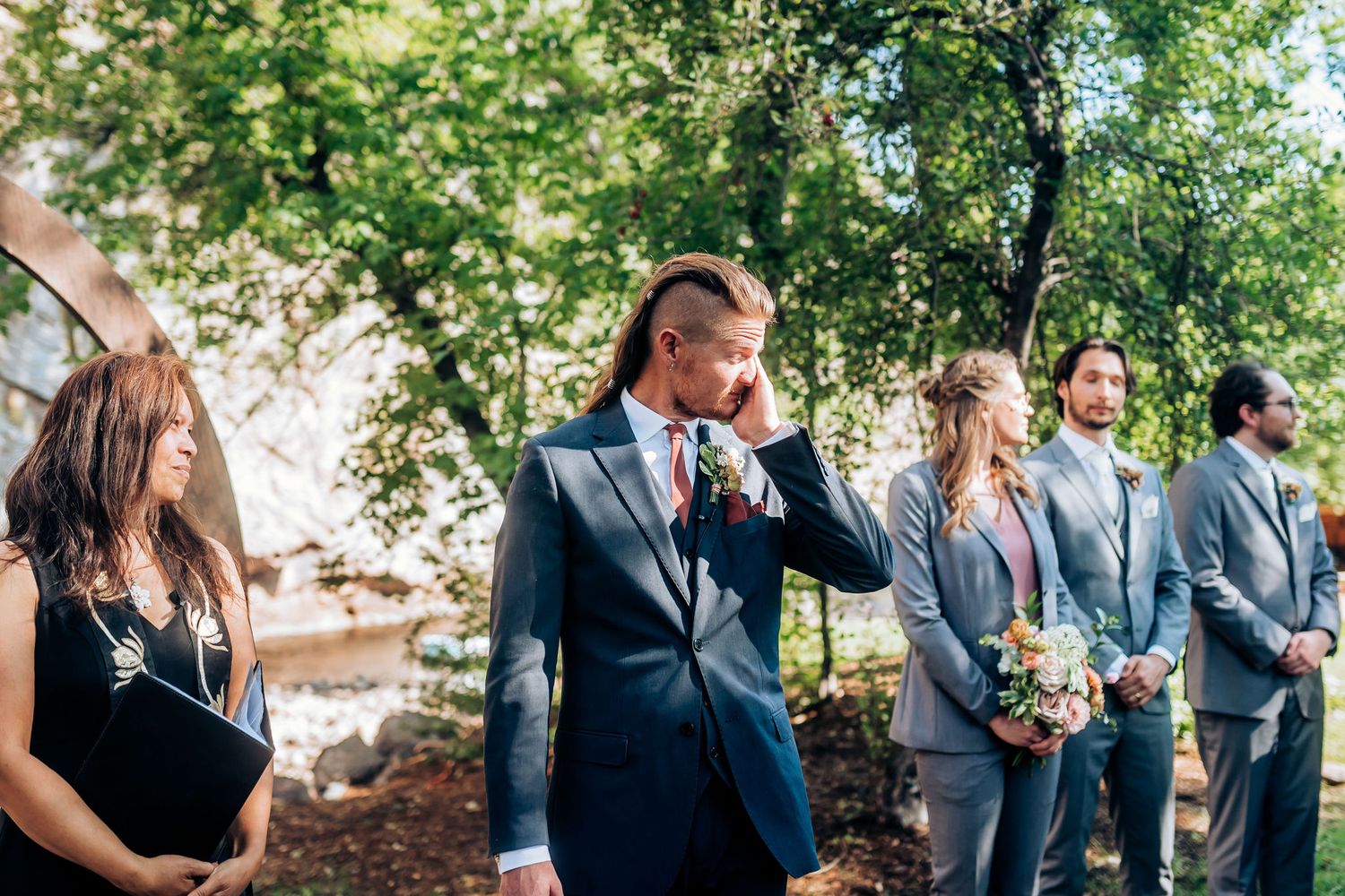 Groomsmen in formal attire standing in line during outdoor wedding ceremony.