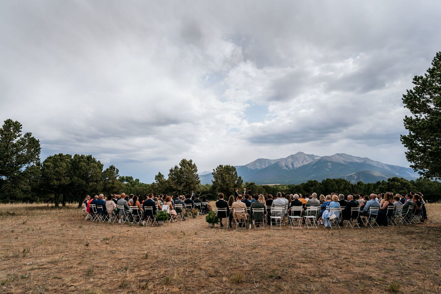 Wedding guests seated in chairs for an outdoor ceremony with dramatic mountain views and cloudy skies.