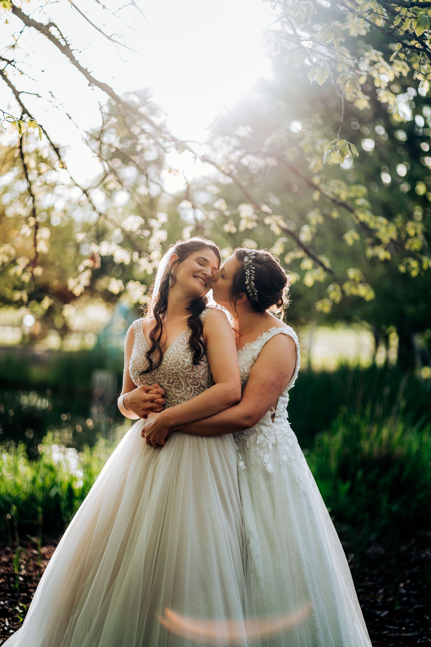 Two brides sharing an intimate moment in white wedding dresses against a sunlit outdoor garden setting.