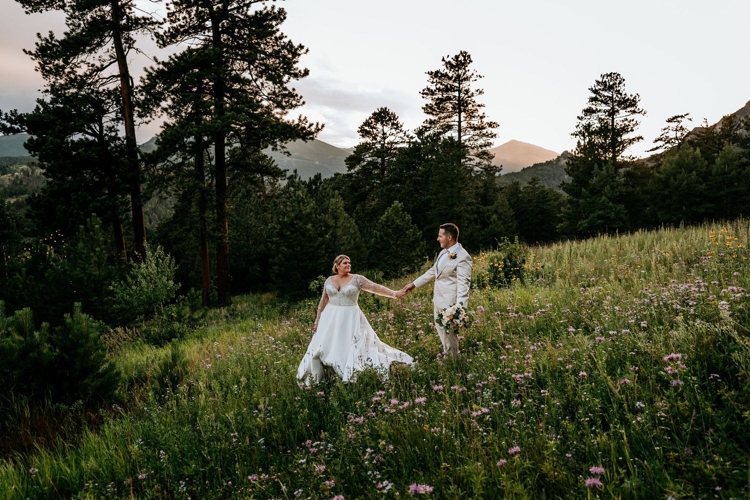 Wedding couple walking hand in hand through a scenic mountain meadow at sunset.